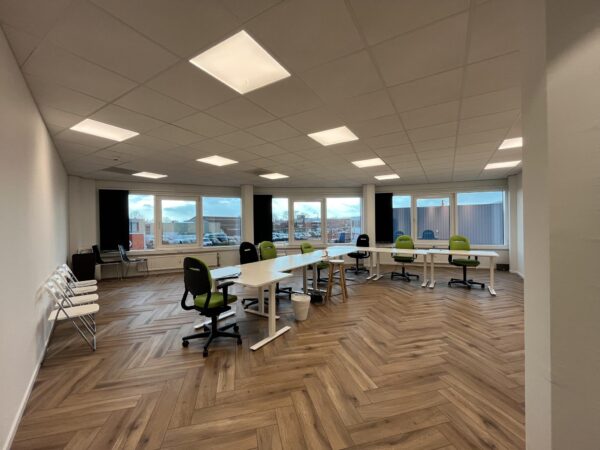 Vergaderruimte aan de Lange Dreef met moderne bureaustoelen, houten visgraatvloer en grote ramen die uitzicht bieden op parkeerplaats en gebouwen.